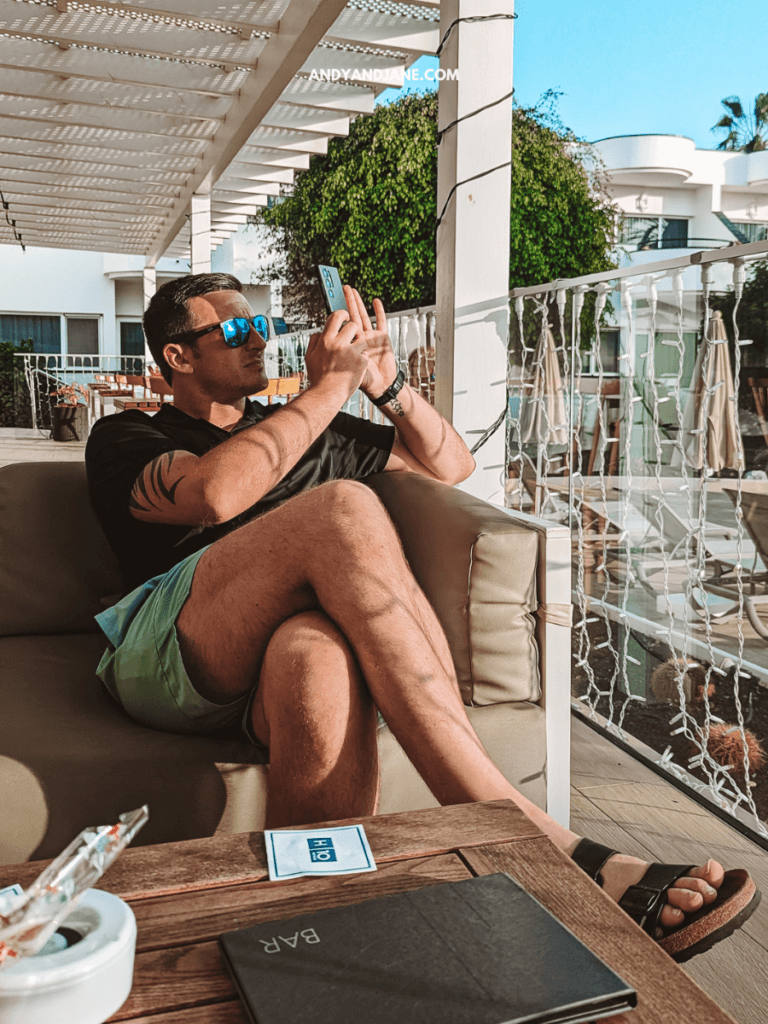 A man sits casually on a couch, taking a photo with his phone, surrounded by a sunny outdoor setting and greenery.