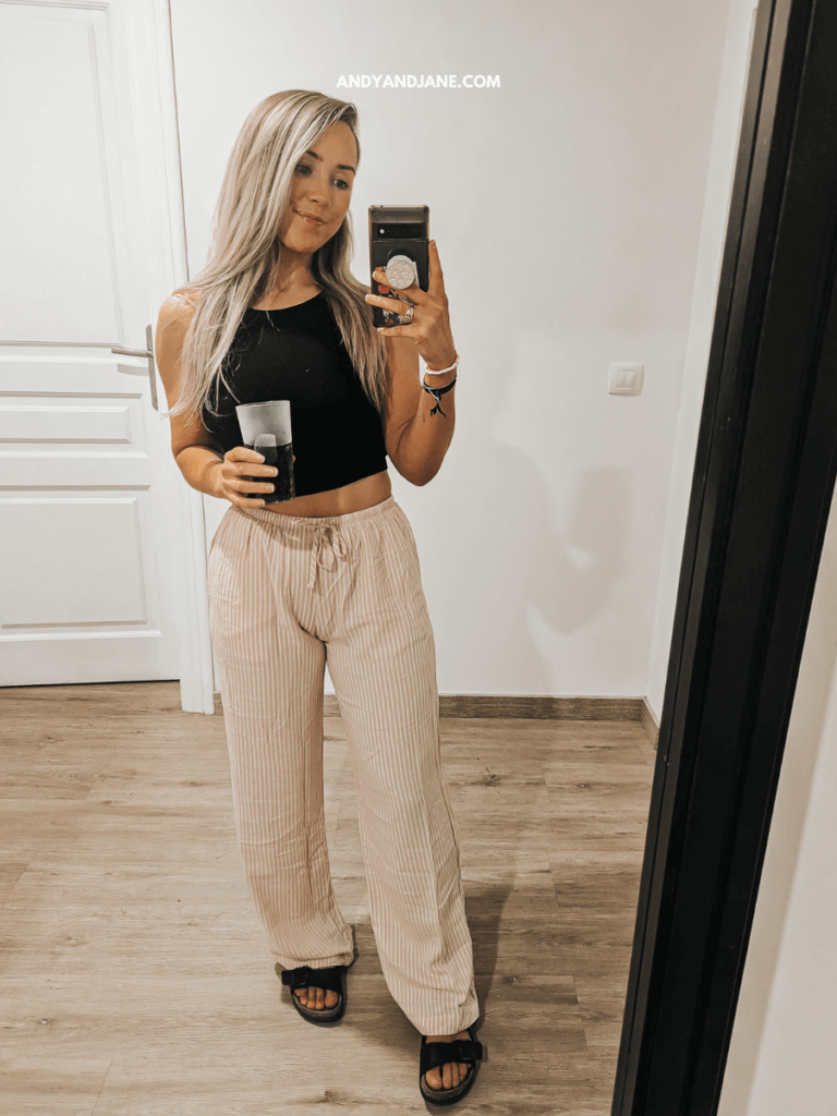 A person stands in a well-lit room holding a glass while wearing a black crop top, striped wide-leg pants, and black sandals.