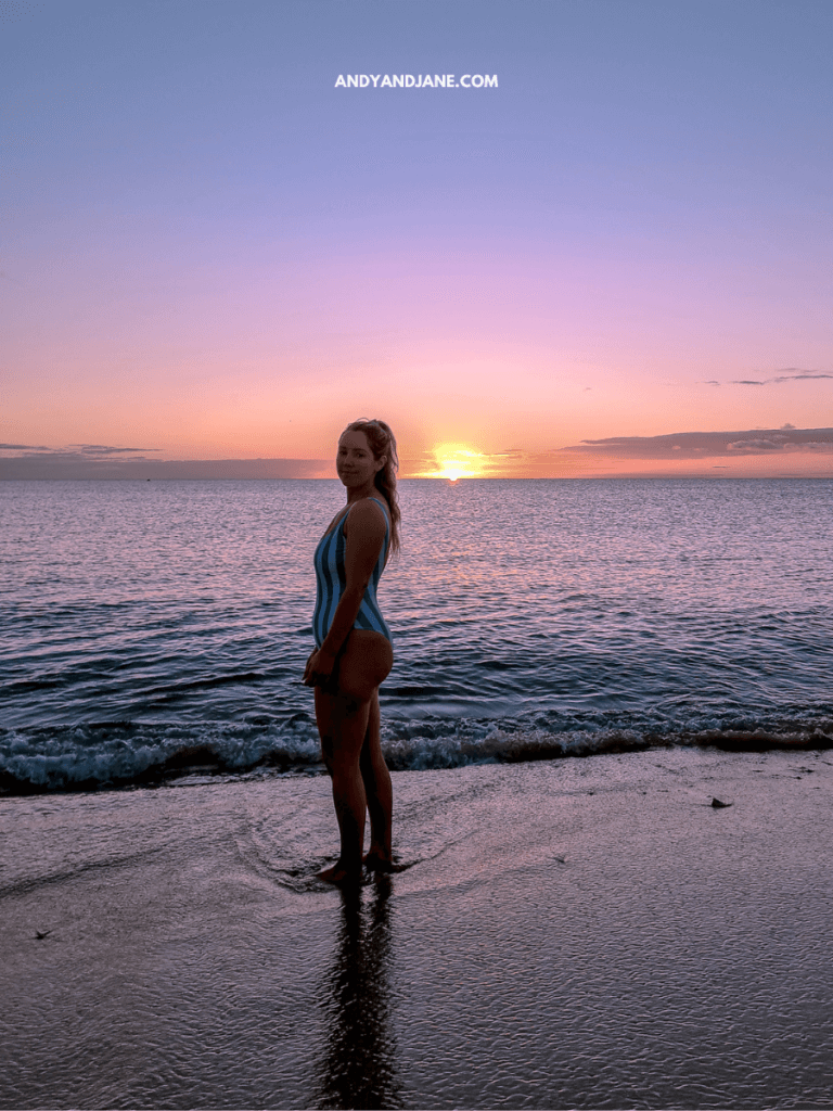 A person in a blue striped swimsuit stands at the shoreline, gazing at a vibrant sunset over the calm ocean waves.