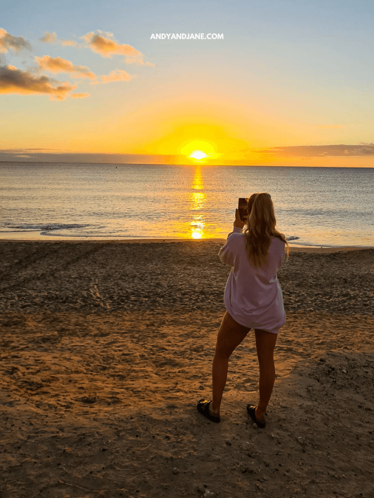 A woman in a white shirt stands on the beach, capturing a vibrant sunset over the calm ocean.