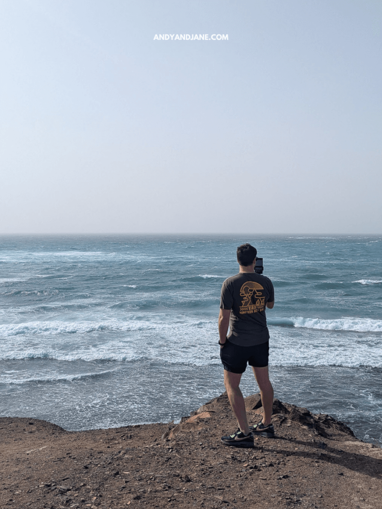 A man in shorts and a t-shirt stands on rocky cliffs, capturing the ocean waves with his phone on a hazy day.