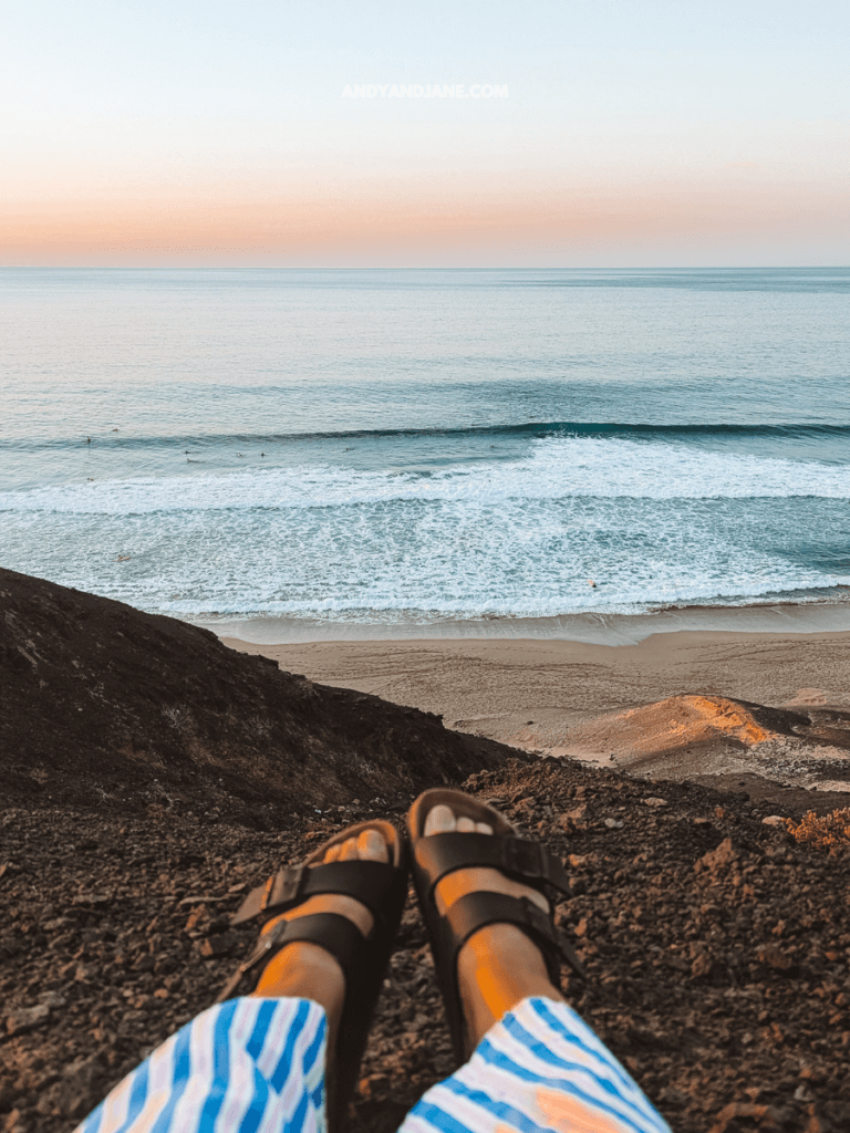 Feet in black sandals atop a rocky cliff, overlooking a serene beach and ocean at sunset, with gentle waves and surfers visible.