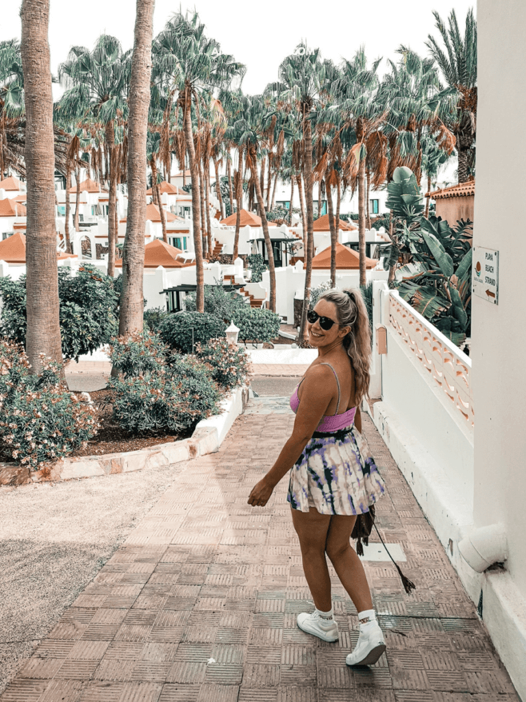 A woman walks down a sunlit pathway lined with palm trees and white buildings with orange roofs, dressed in a colorful skirt and tank top.